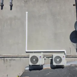 Dwa klimatyzatory marki Gree zamontowane na szarej ścianie budynku z widocznymi rurami odprowadzającymi skropliny i anteną satelitarną w tle.