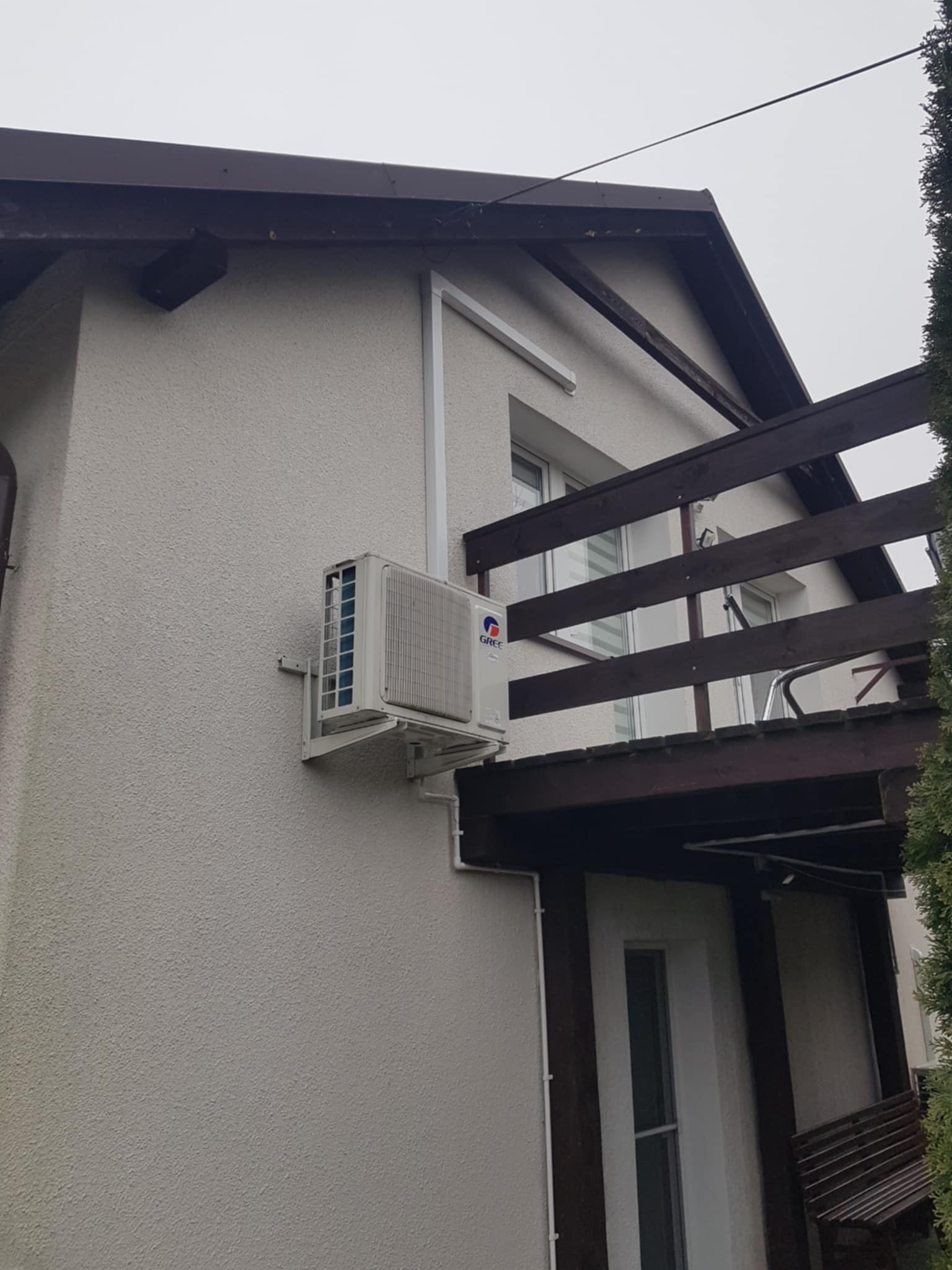 Zewnętrzna jednostka klimatyzacji GREE zamontowana na elewacji domu z fakturą baranka, widoczna drewniana konstrukcja balkonu.