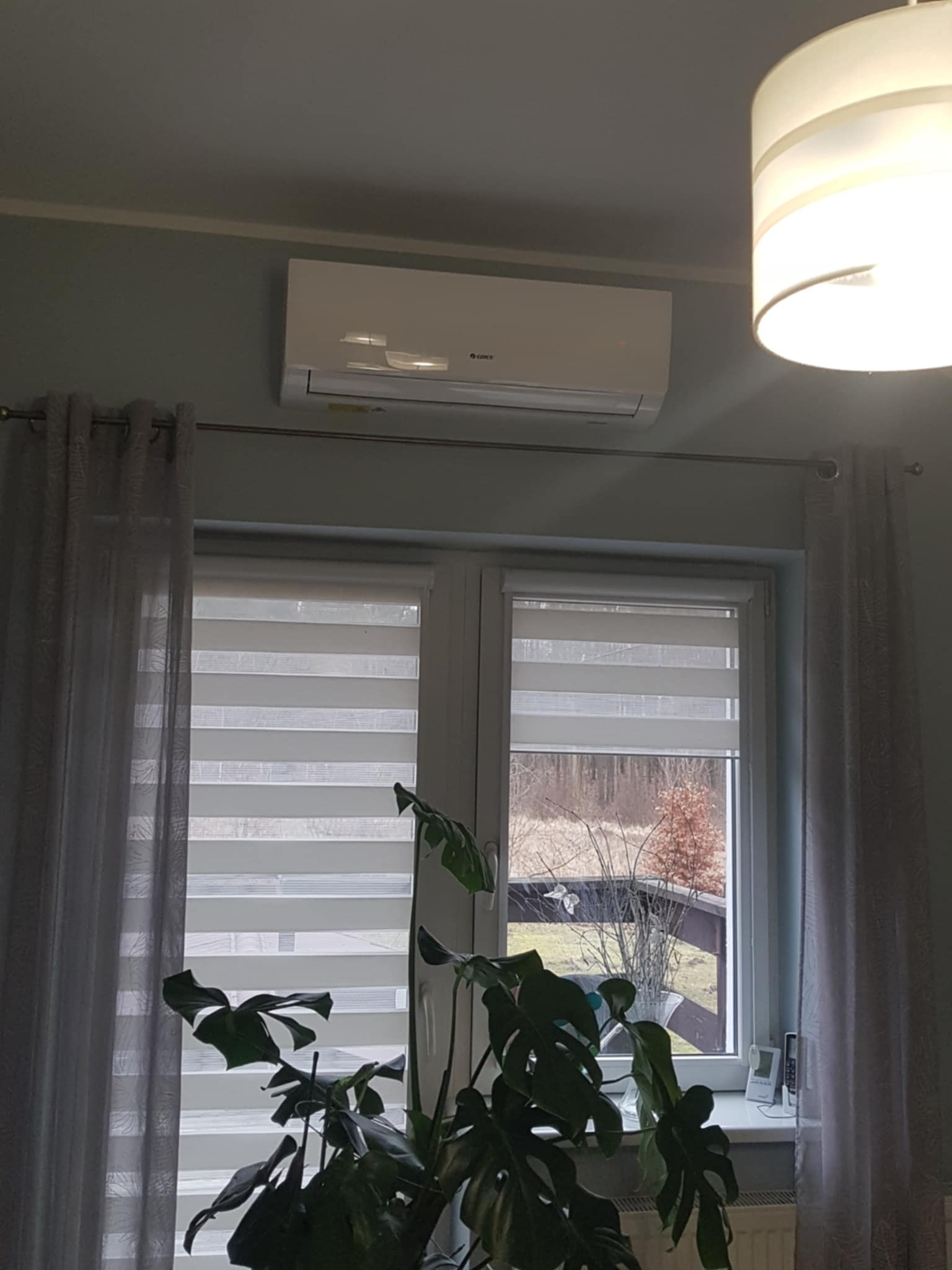 Biały klimatyzator marki Gree zamontowany nad oknem z żaluzjami poziomymi w jasnym pokoju, widok częściowo zasłonięty przez dużą roślinę doniczkową.