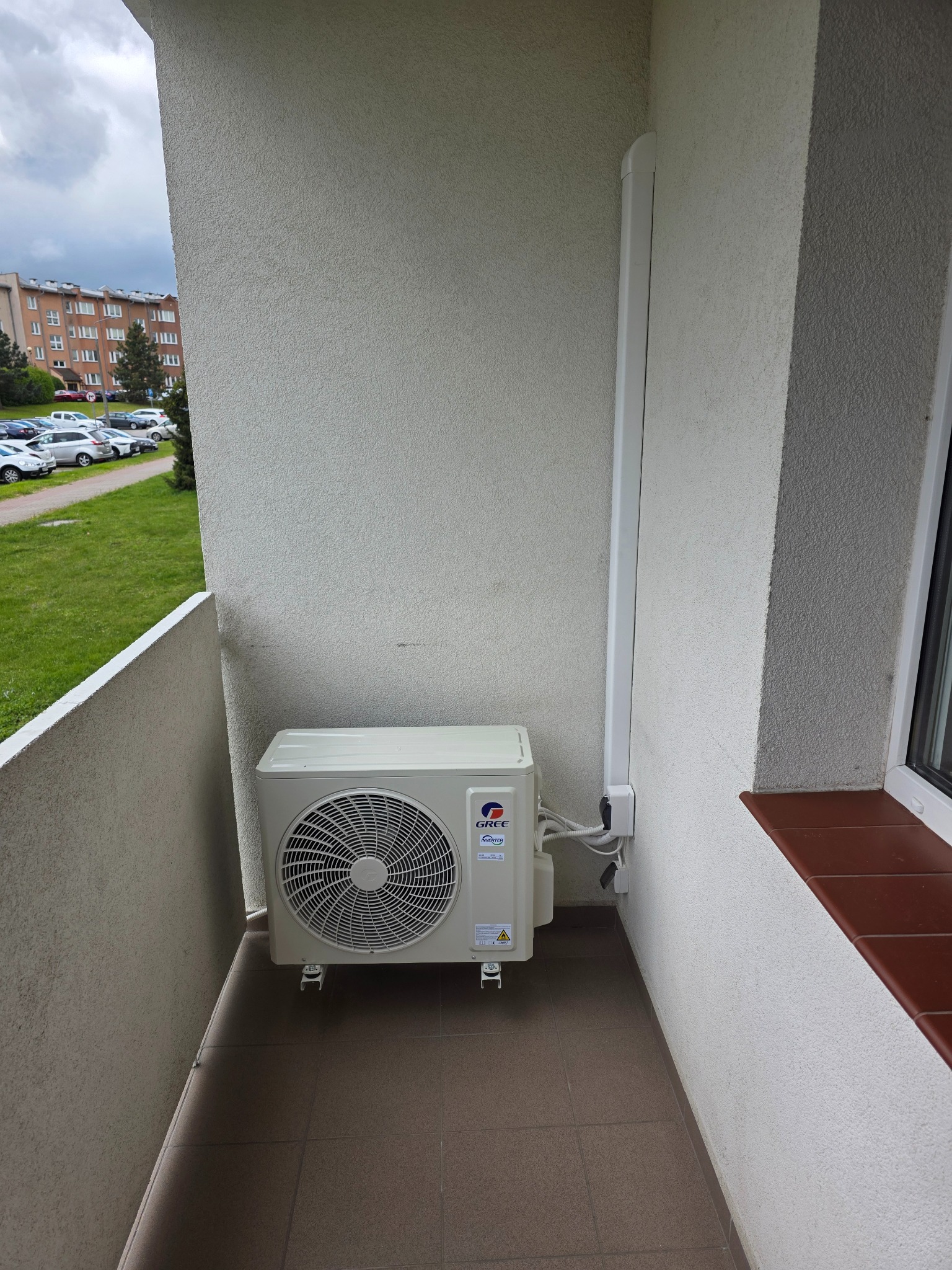 Kompaktowa klimatyzacja Gree na balkonie bloku mieszkalnego, z widoczną listwą maskującą przewody. Zewnętrzny widok na parking i zieleń.