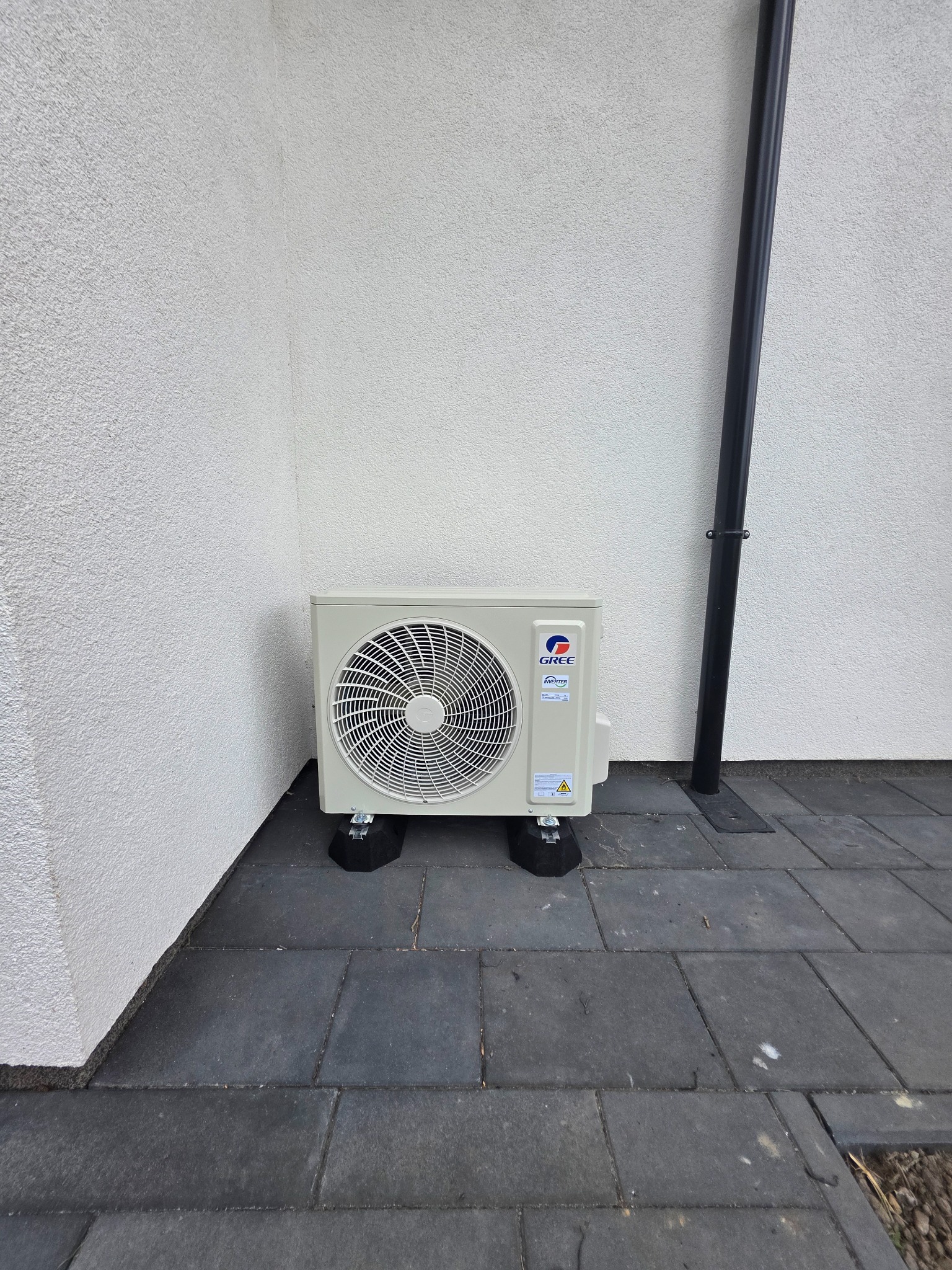 Zewnętrzna jednostka klimatyzacji Gree na czarnych podkładkach antywibracyjnych, ustawiona przy białej ścianie budynku i czarnej rurze spustowej.
