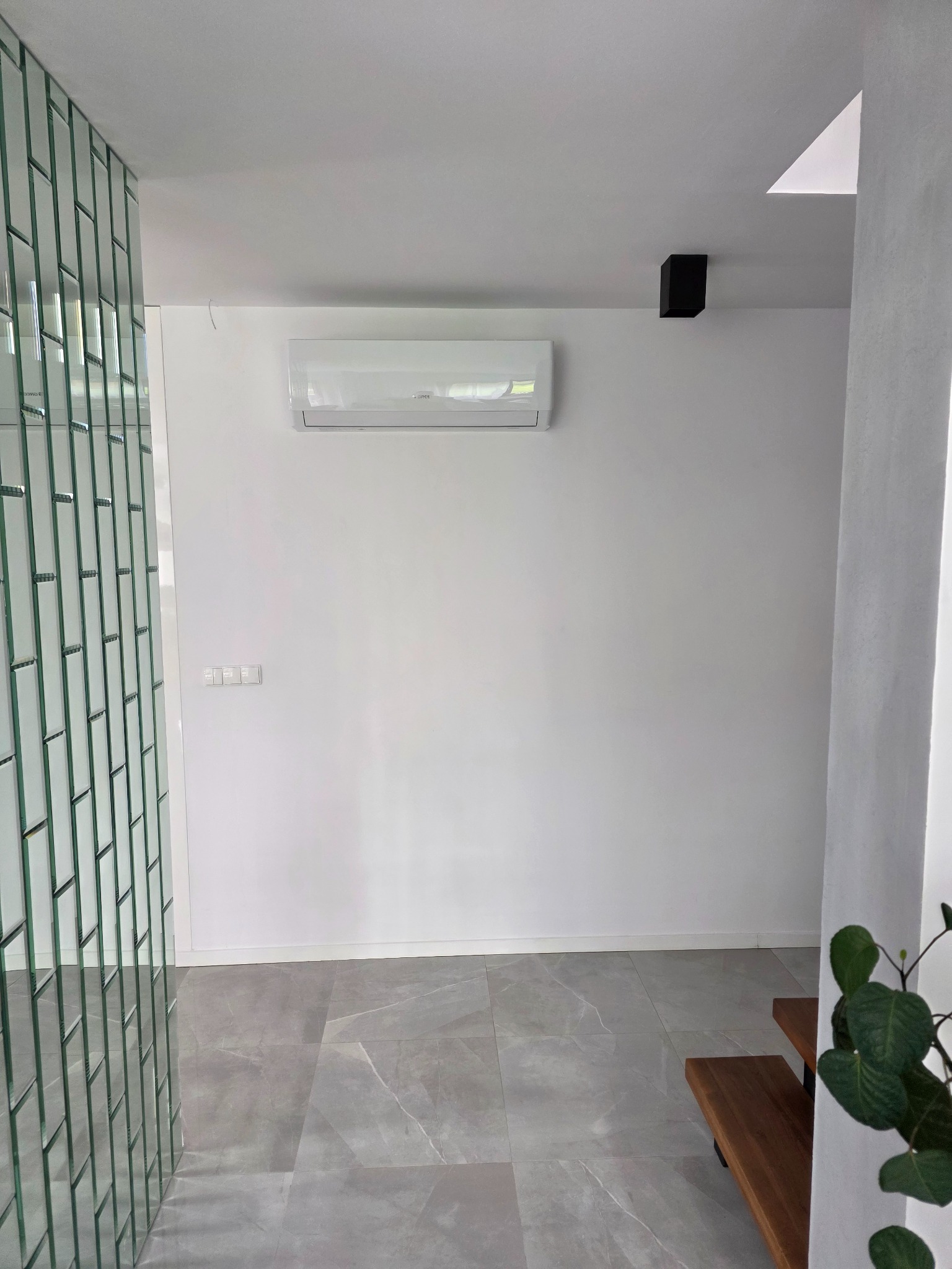 Biały klimatyzator na ścianie w nowoczesnym, minimalistycznym wnętrzu. Szklana ściana z cegiełek i szara podłoga z płytek. Czarny reflektor sufitowy.