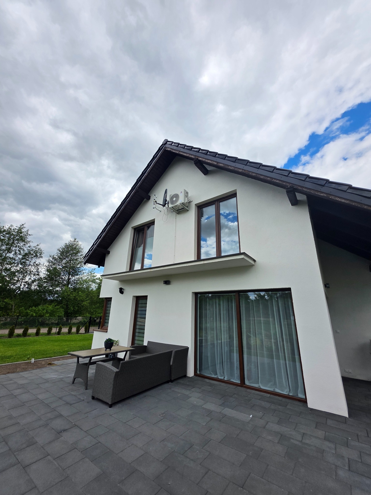 Biały dom z klimatyzacją na elewacji i tarasem z meblami ogrodowymi. W oknach odbija się błękitne niebo. Kątowe ujęcie podkreśla nowoczesny design.