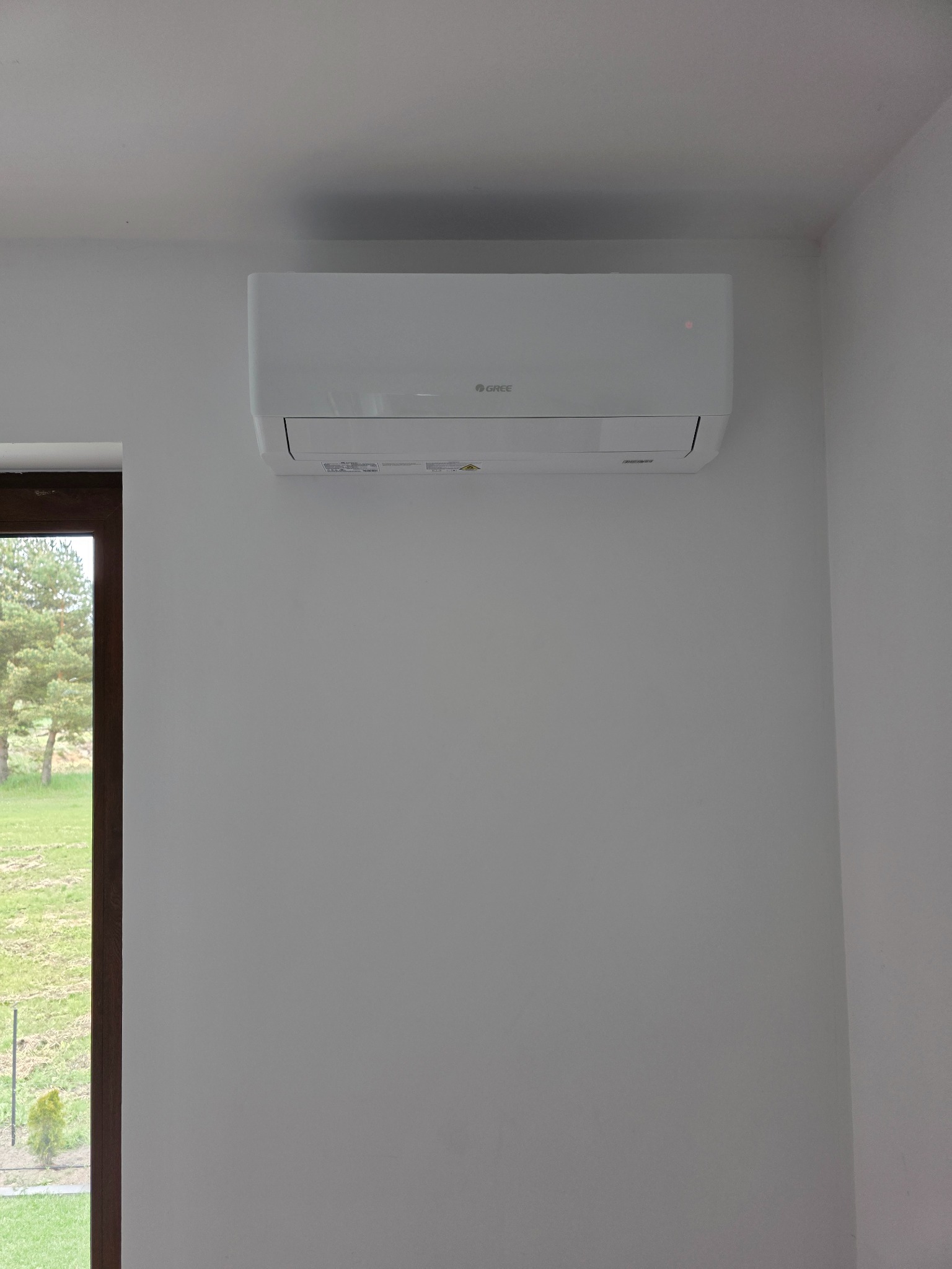 Biały klimatyzator marki Gree zamontowany na białej ścianie w pomieszczeniu z widokiem na zieleń przez okno.