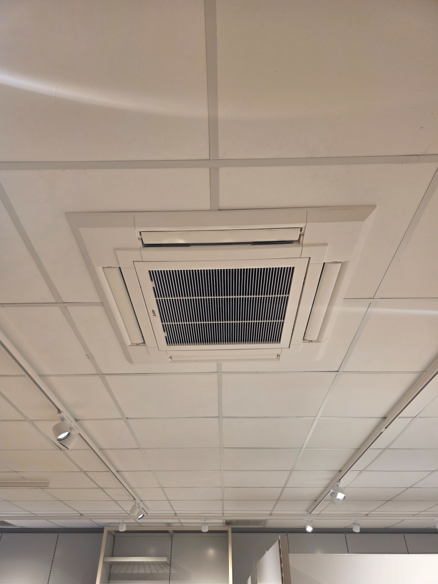 Kasetonowa klimatyzacja sufitowa Mitsubishi Electric, widok z dołu. Biała obudowa, kratka wlotowa. Widoczne oświetlenie szynowe i elementy wyposażenia wnętrza.