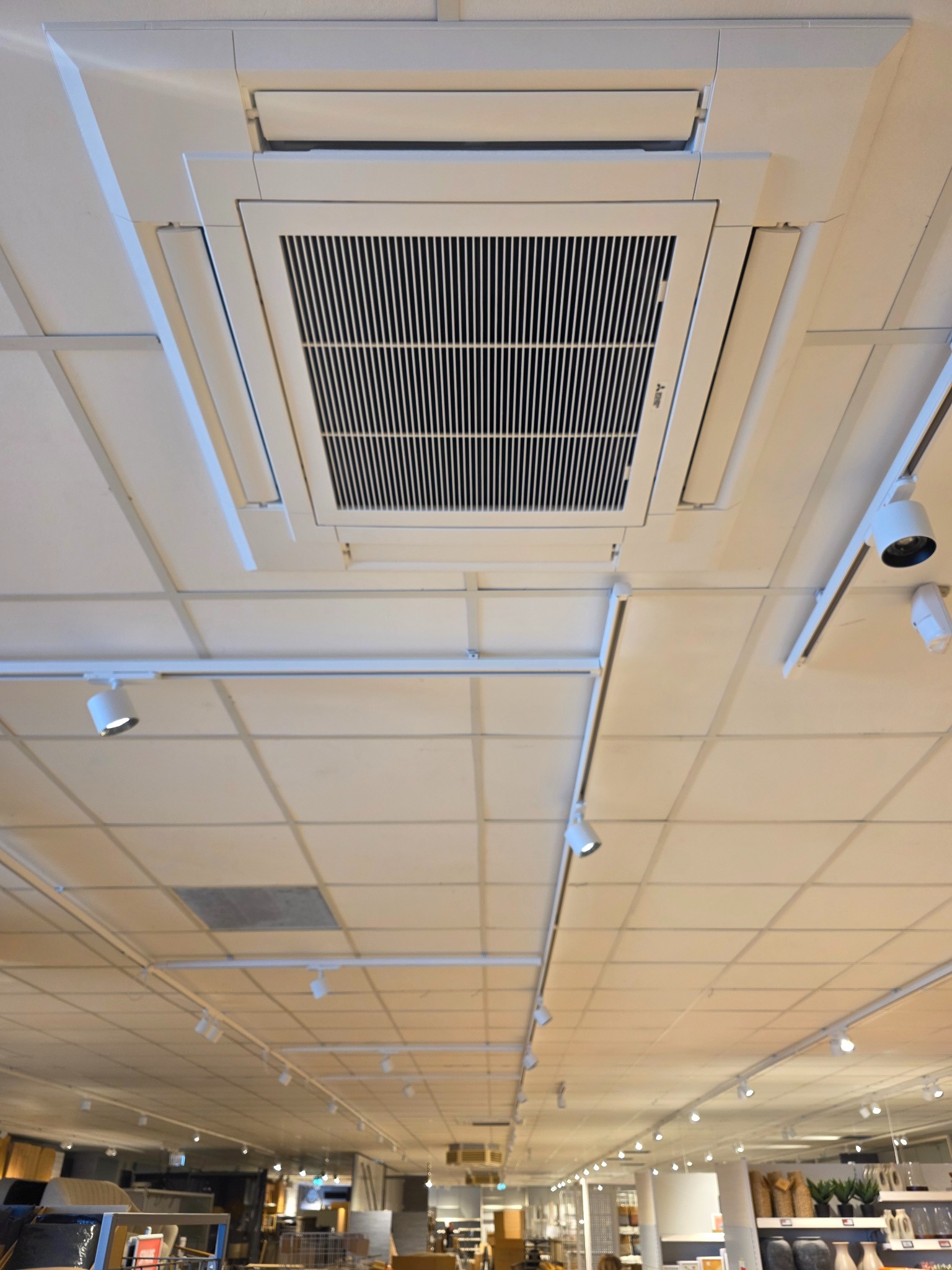 Biała, sufitowa klimatyzacja kasetonowa Mitsubishi Electric, zamontowana w pomieszczeniu z oświetleniem szynowym i podwieszanym sufitem.