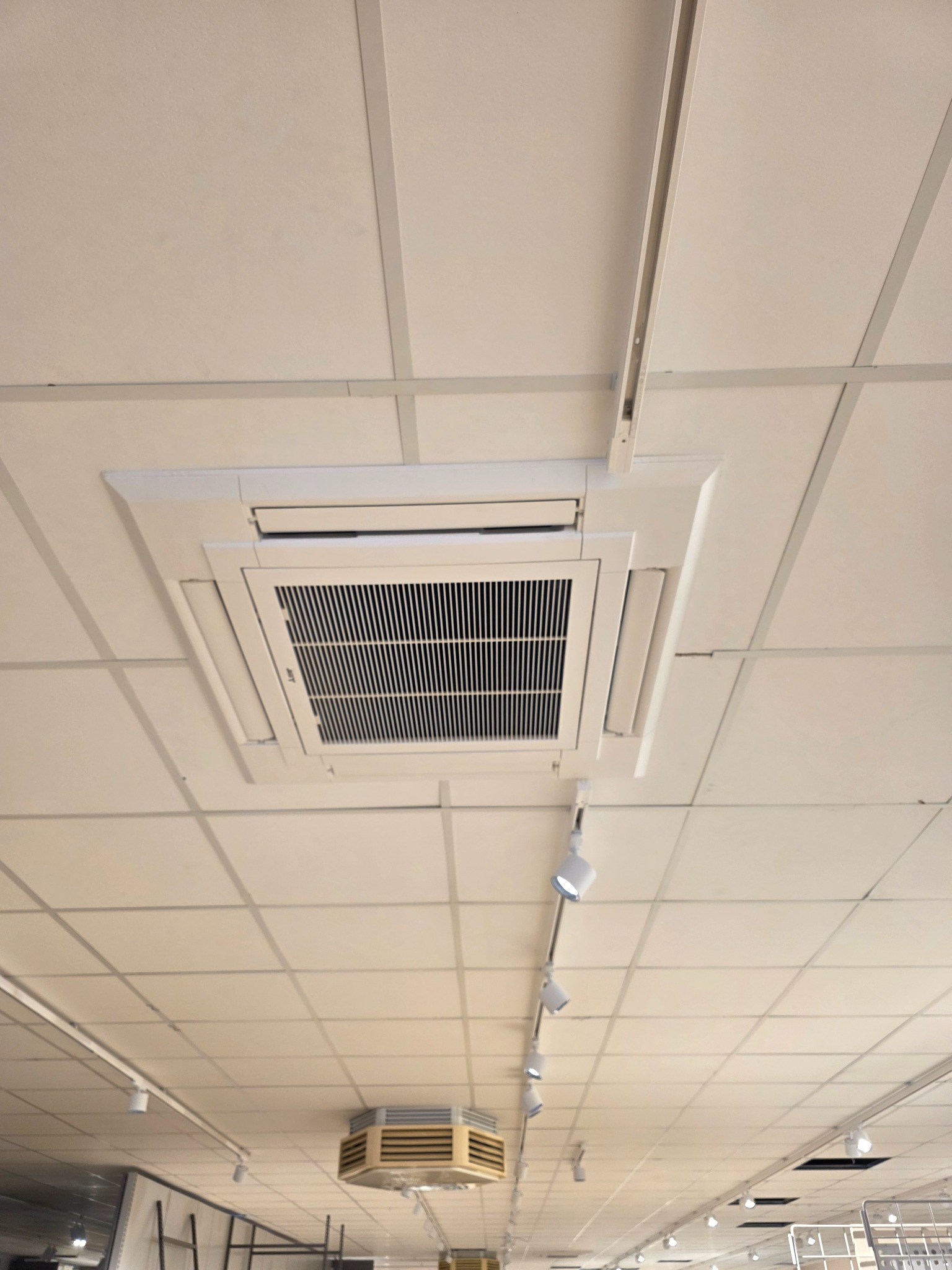 Kasetonowa klimatyzacja sufitowa w biurze z oświetleniem szynowym. Widok z dołu na białą kratkę wentylacyjną i panele sufitowe.