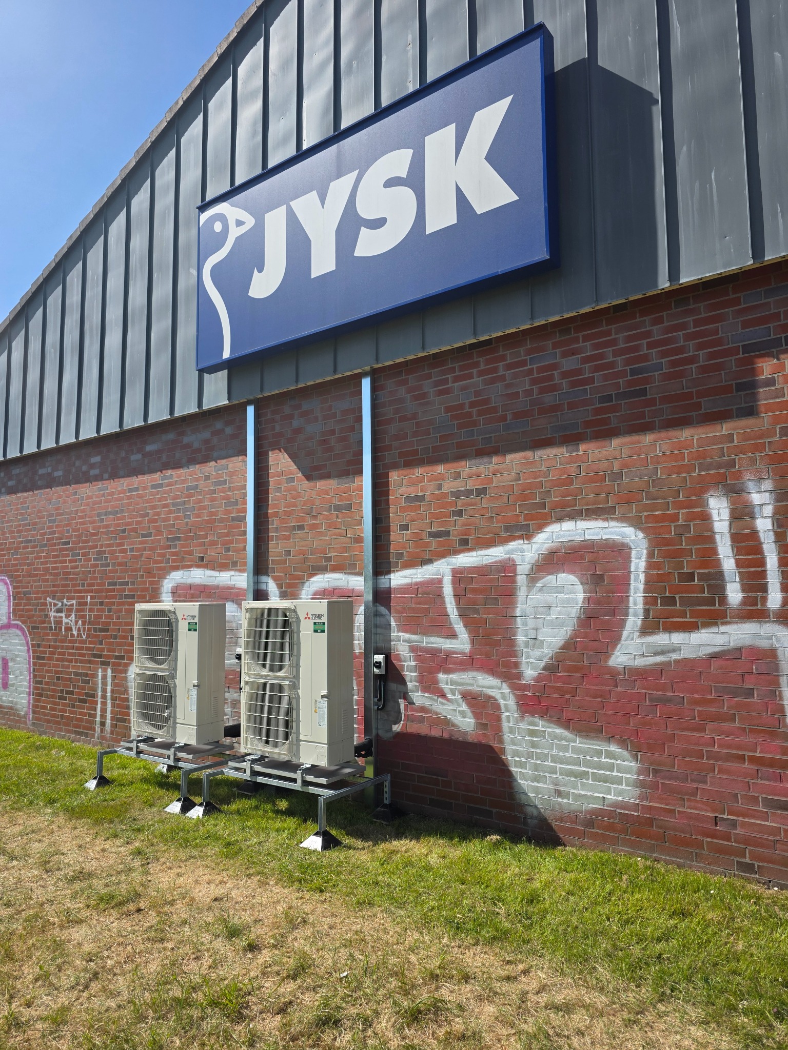 Dwie jednostki klimatyzacji Mitsubishi Electric na metalowej konstrukcji, zamontowane na zewnątrz budynku JYSK z ceglaną ścianą pokrytą graffiti.