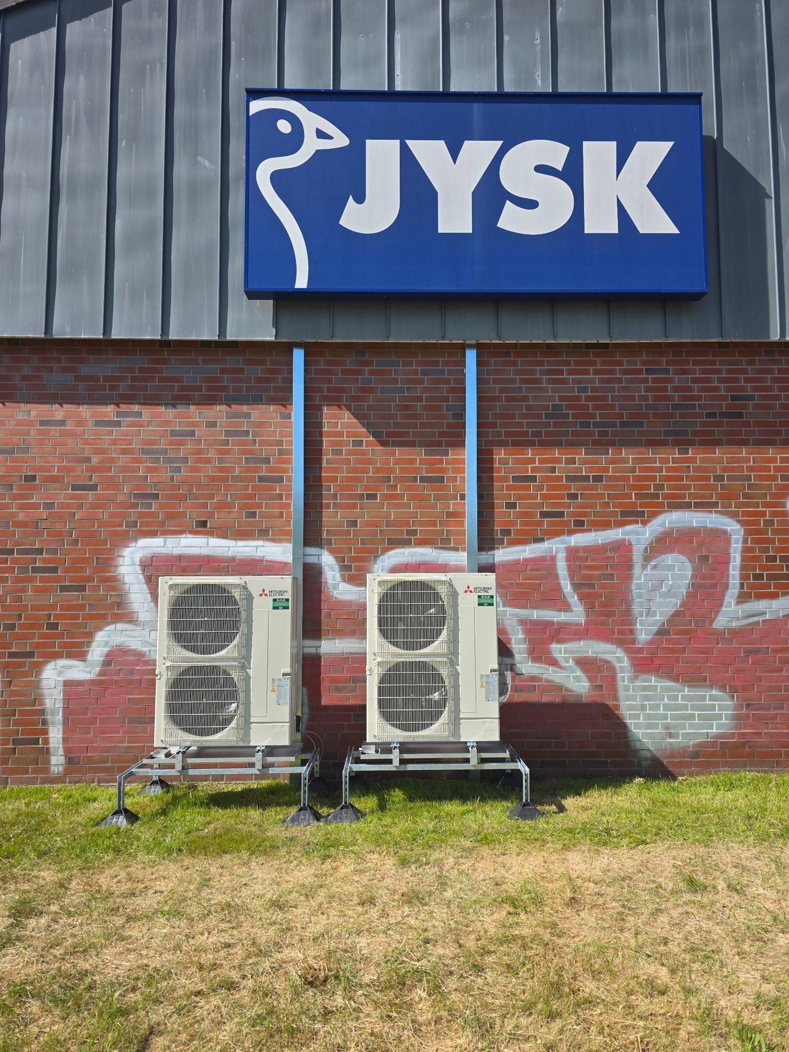 Dwie jednostki zewnętrzne klimatyzacji Mitsubishi na tle ceglanej ściany z graffiti i znaku JYSK. Montaż na trawie, widoczne rury instalacyjne.