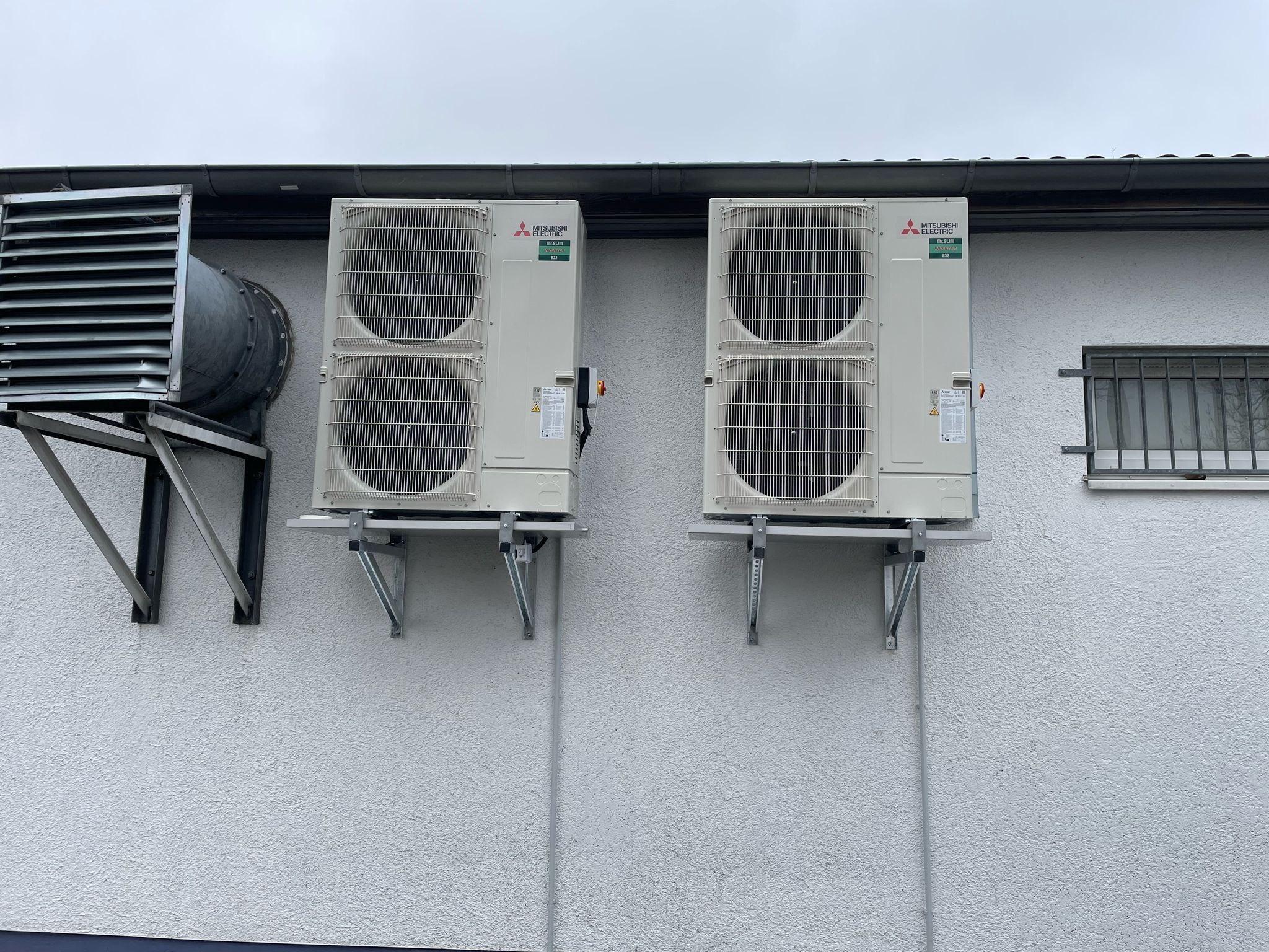 Dwa klimatyzatory Mitsubishi Electric zamontowane na wspornikach na białej, teksturowanej ścianie budynku z szarą rynną i kratką wentylacyjną.