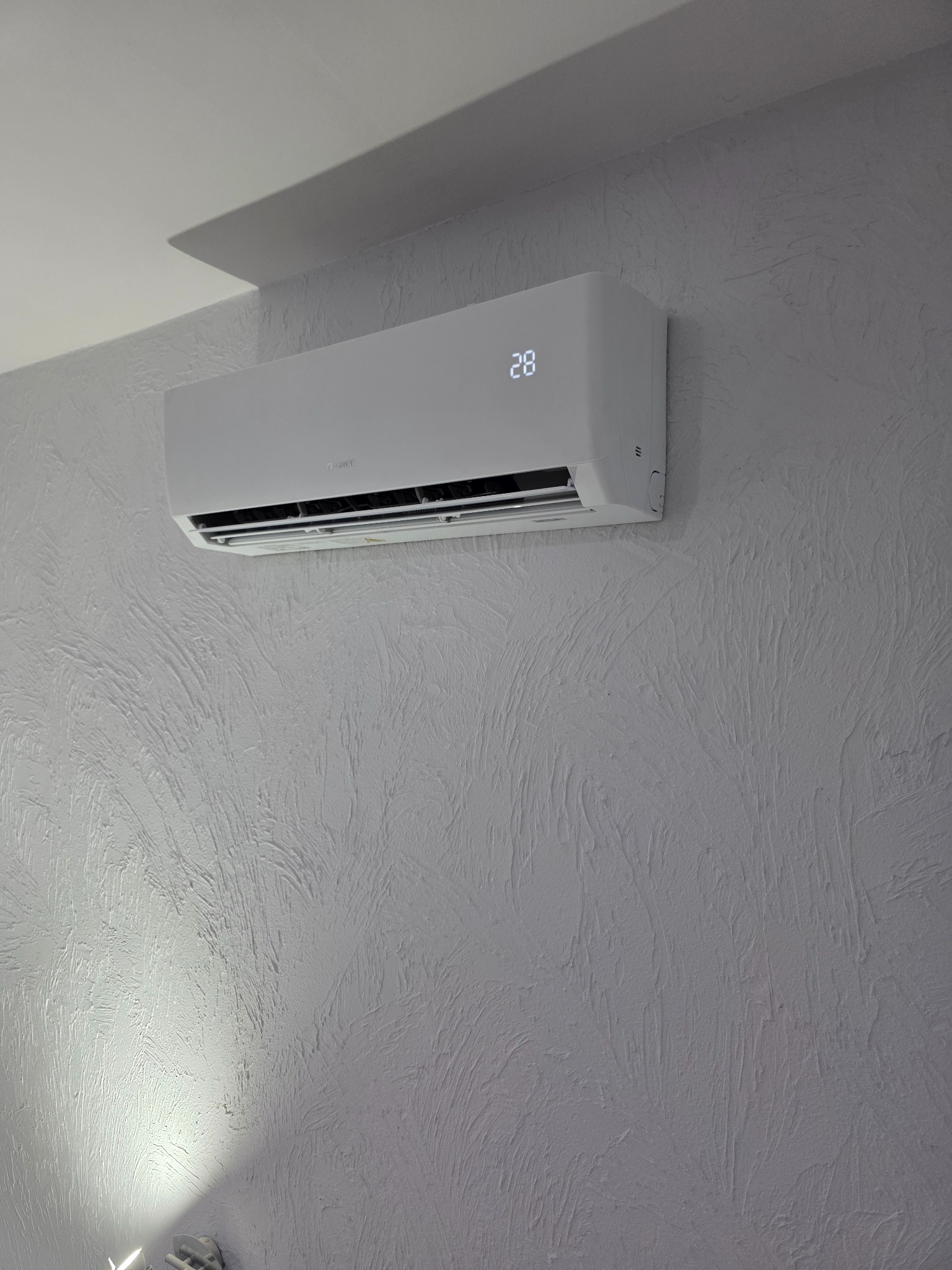 Biały klimatyzator ścienny z wyświetlaczem temperatury 28 stopni na teksturowanej, jasnoszarej ścianie. Widoczny fragment sufitu i oświetlenie.