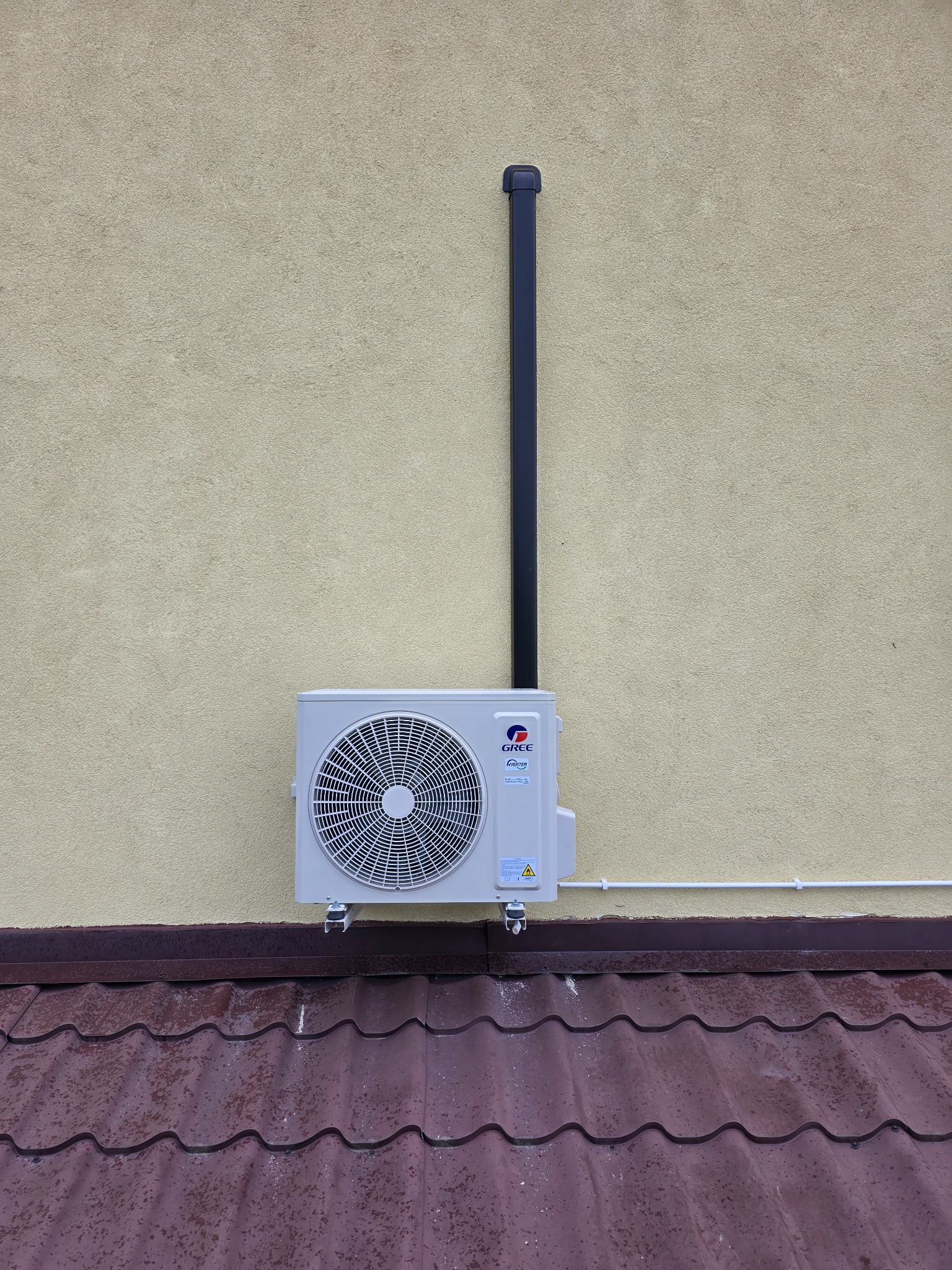 Zewnętrzna jednostka klimatyzacji GREE zamontowana na elewacji budynku z brązowym dachem. Widoczna rura odprowadzająca skropliny i fragment orynnowania.