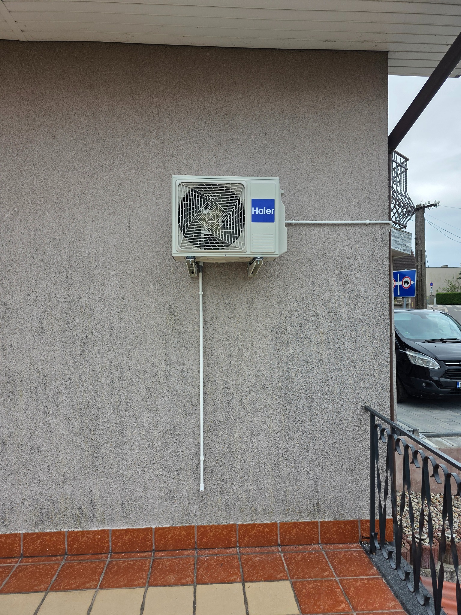 Biała jednostka zewnętrzna klimatyzacji Haier zamontowana na szarej, teksturowanej ścianie budynku z widocznymi rurami i elementami instalacji.