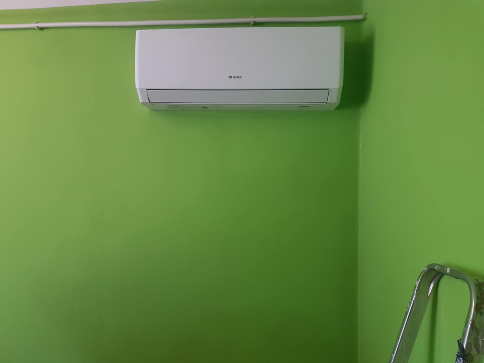 Biały klimatyzator marki Gree zamontowany na intensywnie zielonej ścianie. Fragment drabiny w prawym dolnym rogu. Widoczna rura instalacyjna pod sufitem.