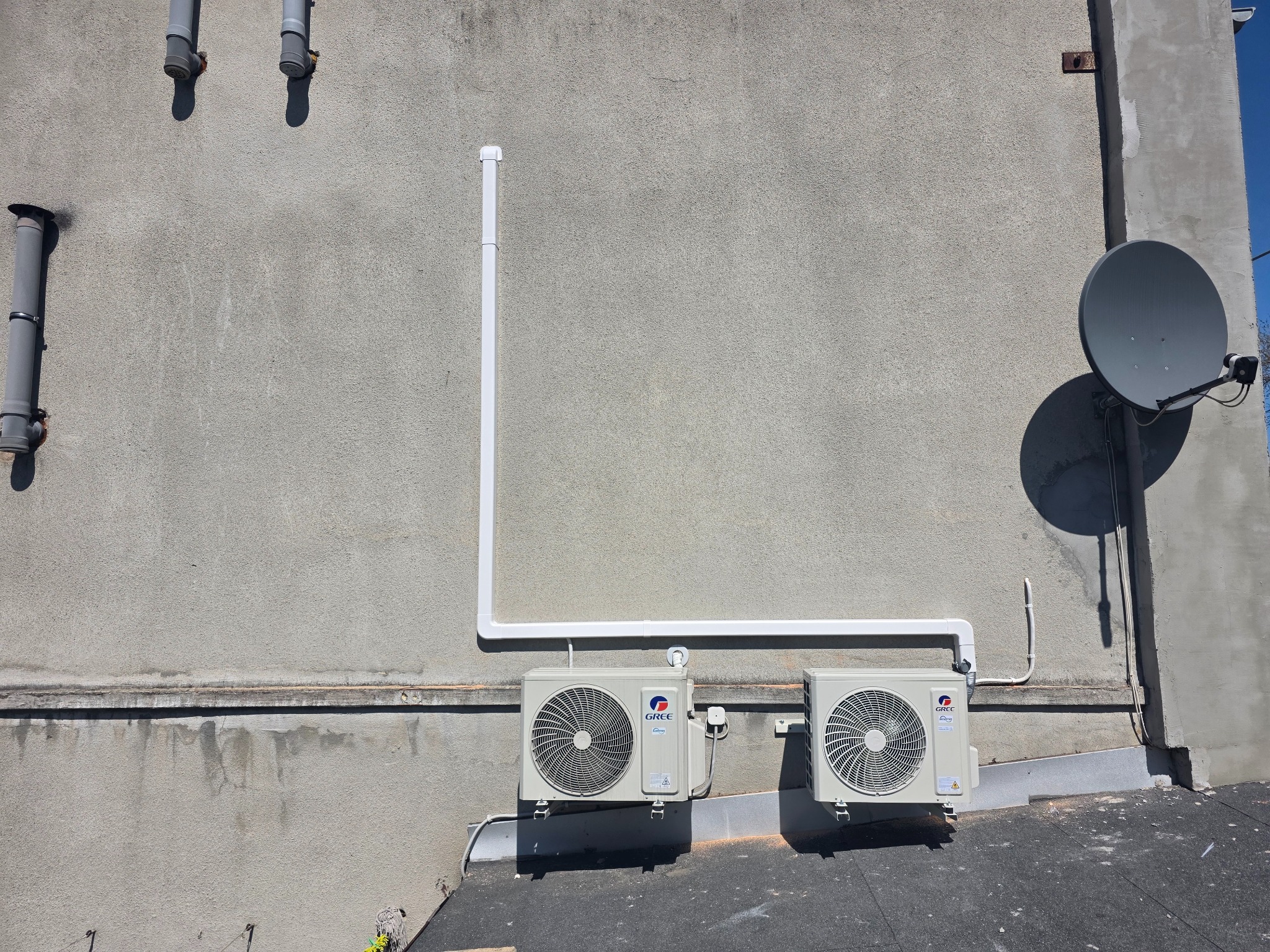 Dwa klimatyzatory marki Gree zamontowane na szarej ścianie budynku z widocznymi rurami odprowadzającymi skropliny i anteną satelitarną w tle.