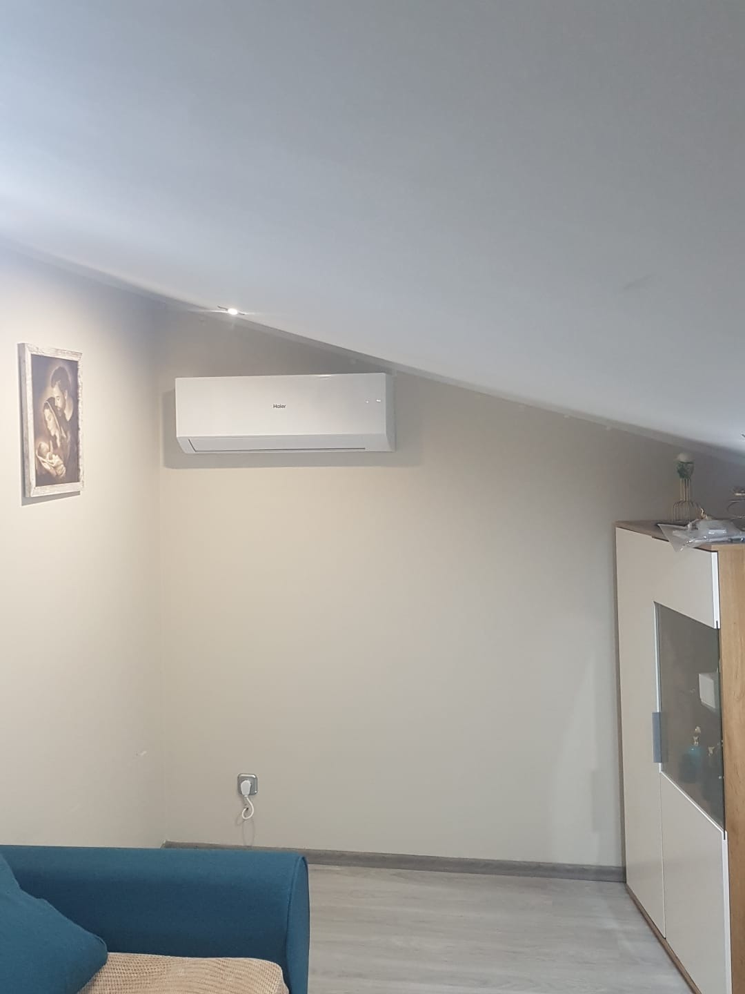 Biała klimatyzacja Haier zamontowana na ścianie w pokoju ze skośnym sufitem. Widoczny fragment niebieskiej sofy, obraz na ścianie i biała szafka.