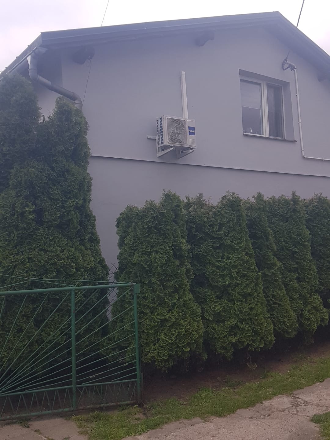 Szary dom jednorodzinny z zamontowanym klimatyzatorem Haier na elewacji, widok z poziomu gruntu, zielone tuje wzdłuż ogrodzenia.