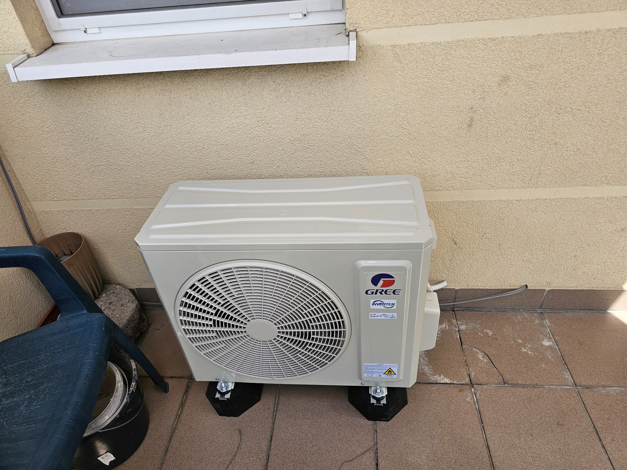 Zewnętrzna jednostka klimatyzacji marki Gree, umieszczona na czarnych podkładkach antywibracyjnych, stoi na brązowych płytkach tarasowych przy beżowej ścianie.