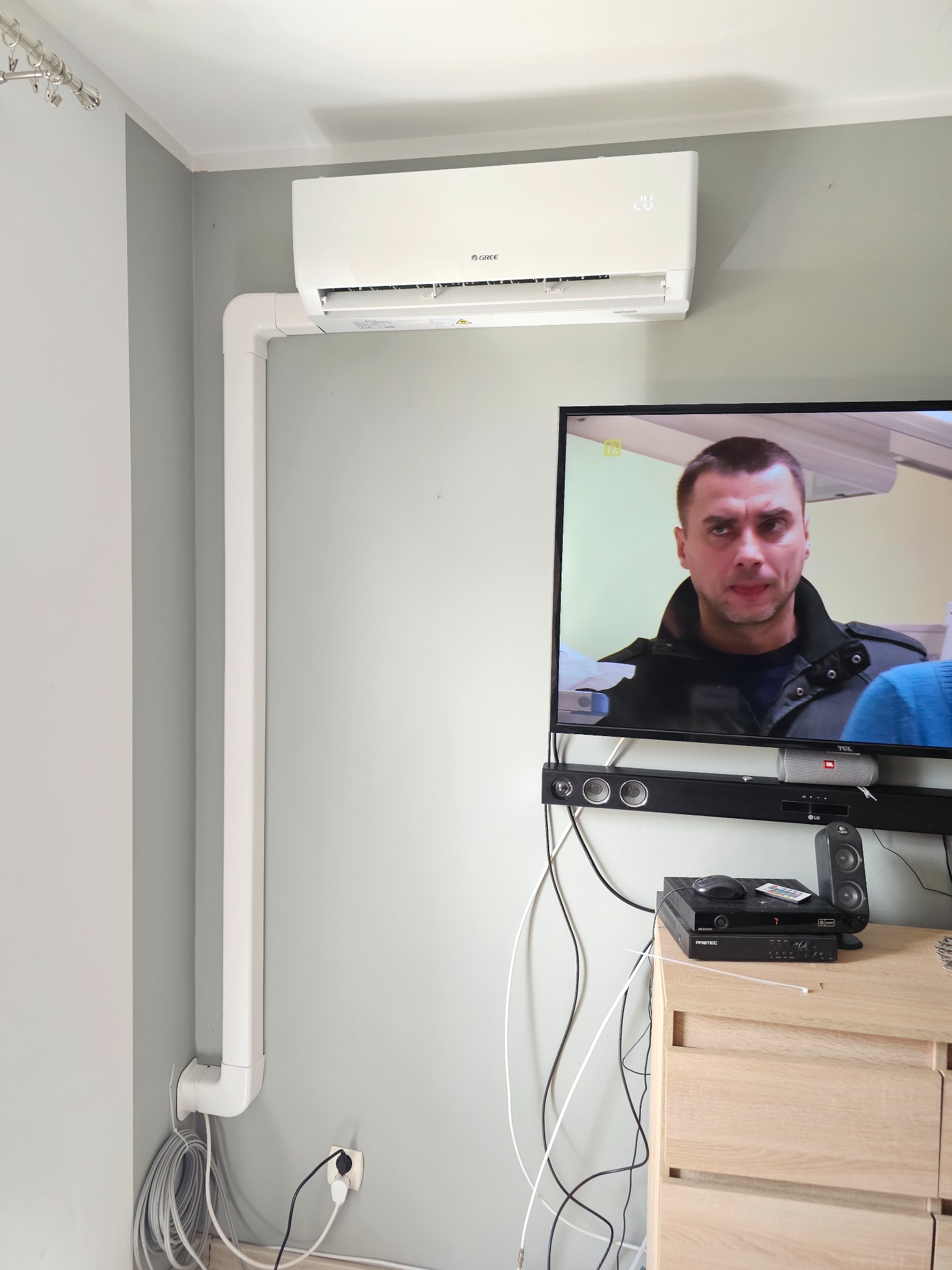 Biała klimatyzacja Gree na ścianie w pokoju, z widocznymi rurami instalacyjnymi, telewizorem i szafką RTV. Ustawiona temperatura 20 stopni.