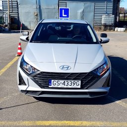AMBER Ośrodek Szkolenia Kierowców - Szary samochód Hyundai z niebieską tablicą 'L' na dachu, zaparkowany na placu manewrowym z żółtymi liniami i czerwono-białymi pachołkami, widziany z przodu, z widoczną osobą za kierownicą.
