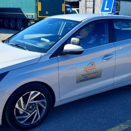AMBER Ośrodek Szkolenia Kierowców - Jasnoszary samochód z naklejką szkoły nauki jazdy 'Amber' na drzwiach, zaparkowany na placu manewrowym z widoczną osobą za kierownicą i znakiem 'L' na dachu.