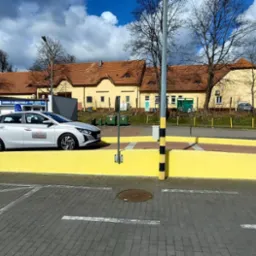 Srebrny samochód z naklejką szkoły jazdy na placu manewrowym z żółtą platformą, białymi liniami na kostce brukowej i budynkami w tle.