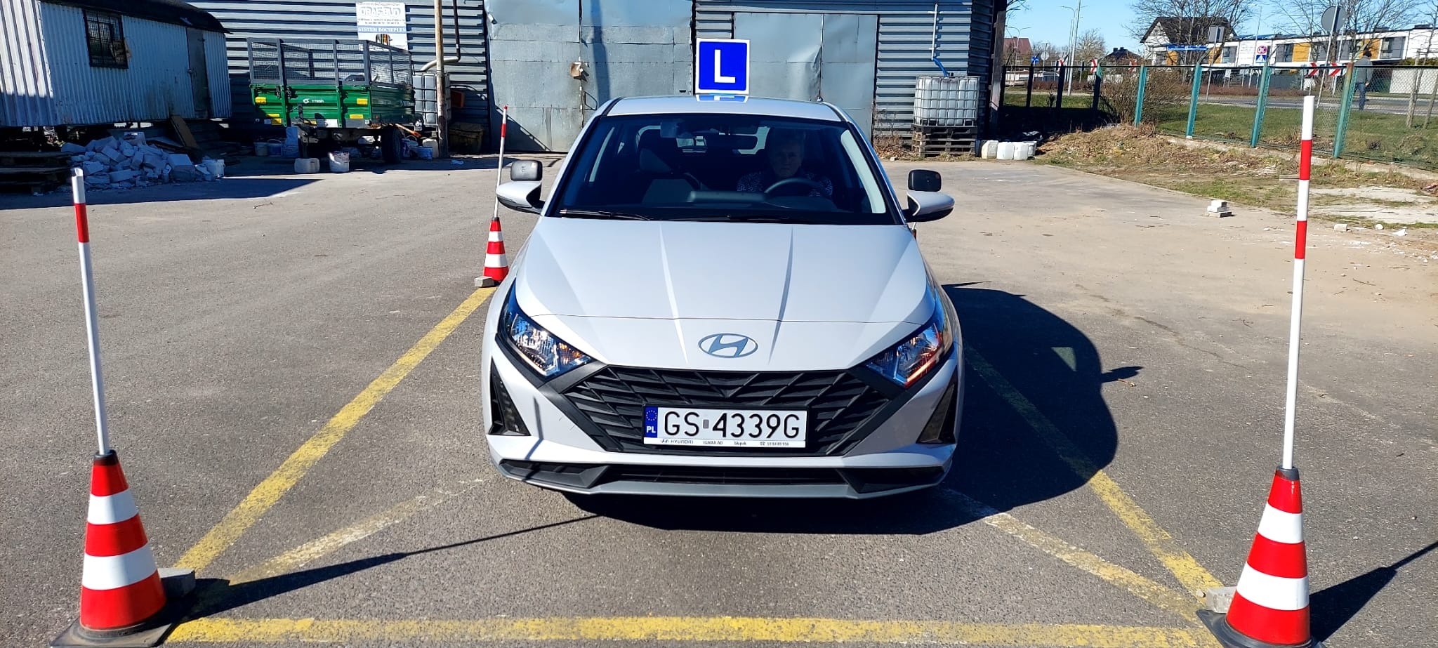 Szary samochód Hyundai z niebieską tablicą 'L' na dachu, zaparkowany na placu manewrowym z żółtymi liniami i czerwono-białymi pachołkami, widziany z przodu, z widoczną osobą za kierownicą.