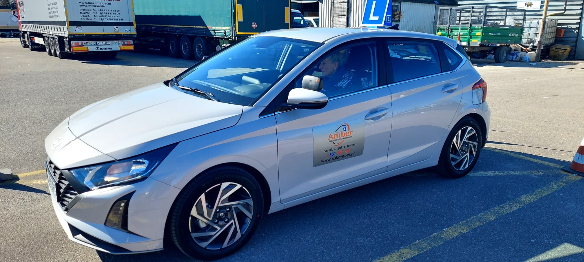 Jasnoszary samochód z naklejką szkoły nauki jazdy 'Amber' na drzwiach, zaparkowany na placu manewrowym z widoczną osobą za kierownicą i znakiem 'L' na dachu.