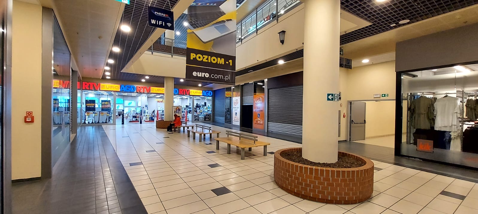 Wnętrze pasażu handlowego z widocznymi sklepami RTV Euro AGD, osobą siedzącą na ławce i szyldem 'POZIOM-1 euro.com.pl'.