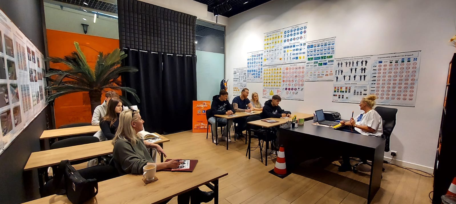 Sala wykładowa szkoły jazdy z grupą uczniów i instruktorką, na ścianach plakaty z przepisami ruchu drogowego, widoczna palma dekoracyjna i stożek ostrzegawczy.