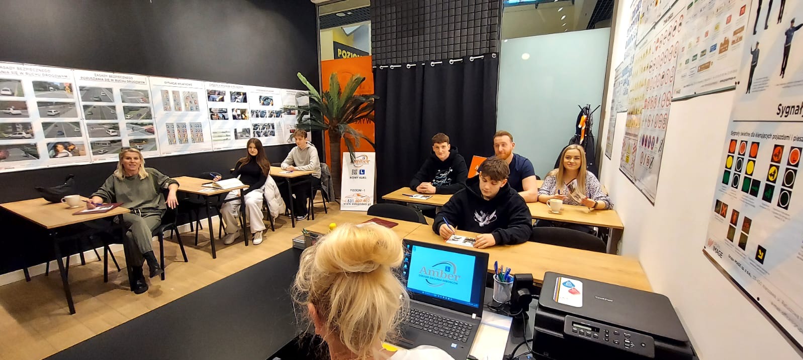 Sala szkoleniowa z uczniami podczas lekcji w szkole jazdy, na ścianach plakaty z zasadami ruchu drogowego i sygnalizacją świetlną, na biurku laptop z logo szkoły.