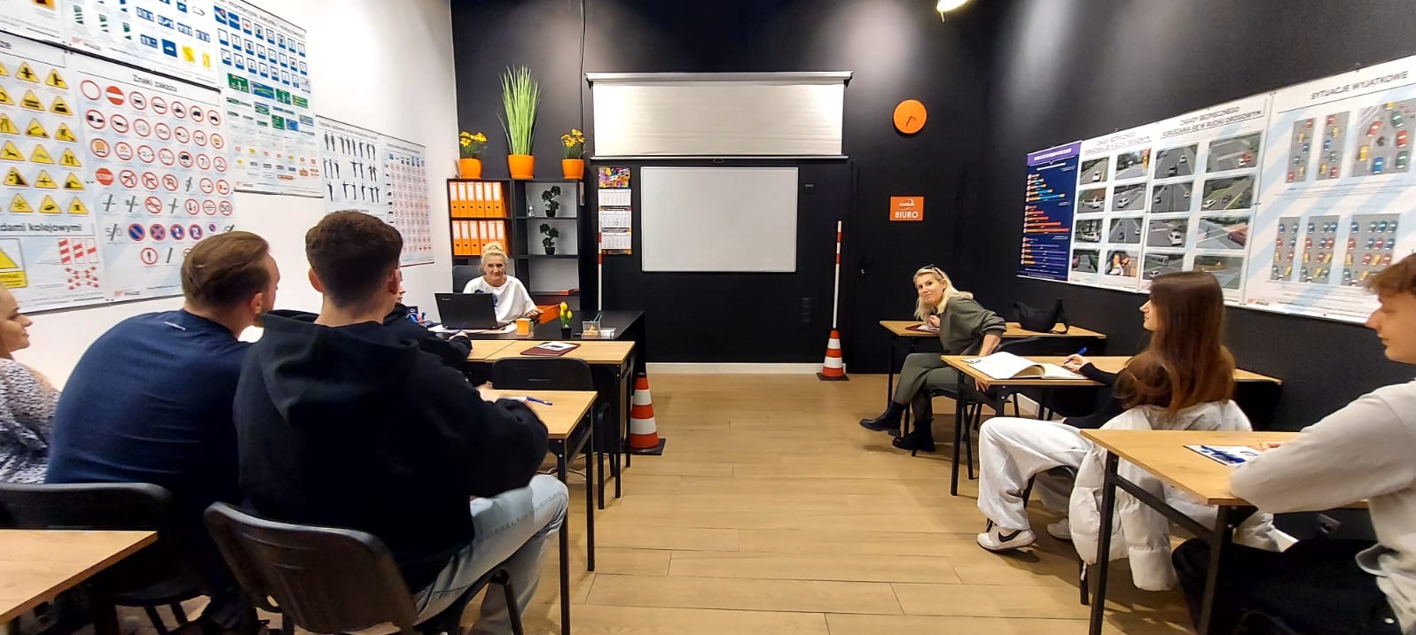 Sala wykładowa z kursantami i instruktorką. Na ścianach plakaty ze znakami drogowymi i sytuacjami drogowymi. W tle biurko z laptopem i pomarańczowymi segregatorami.
