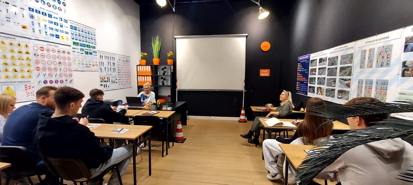 Sala wykładowa z kursantami, instruktorem przy biurku z laptopem, planszami edukacyjnymi dotyczącymi przepisów ruchu drogowego na ścianach, stożkiem ostrzegawczym i rzutnikiem z rozwiniętym ekranem.