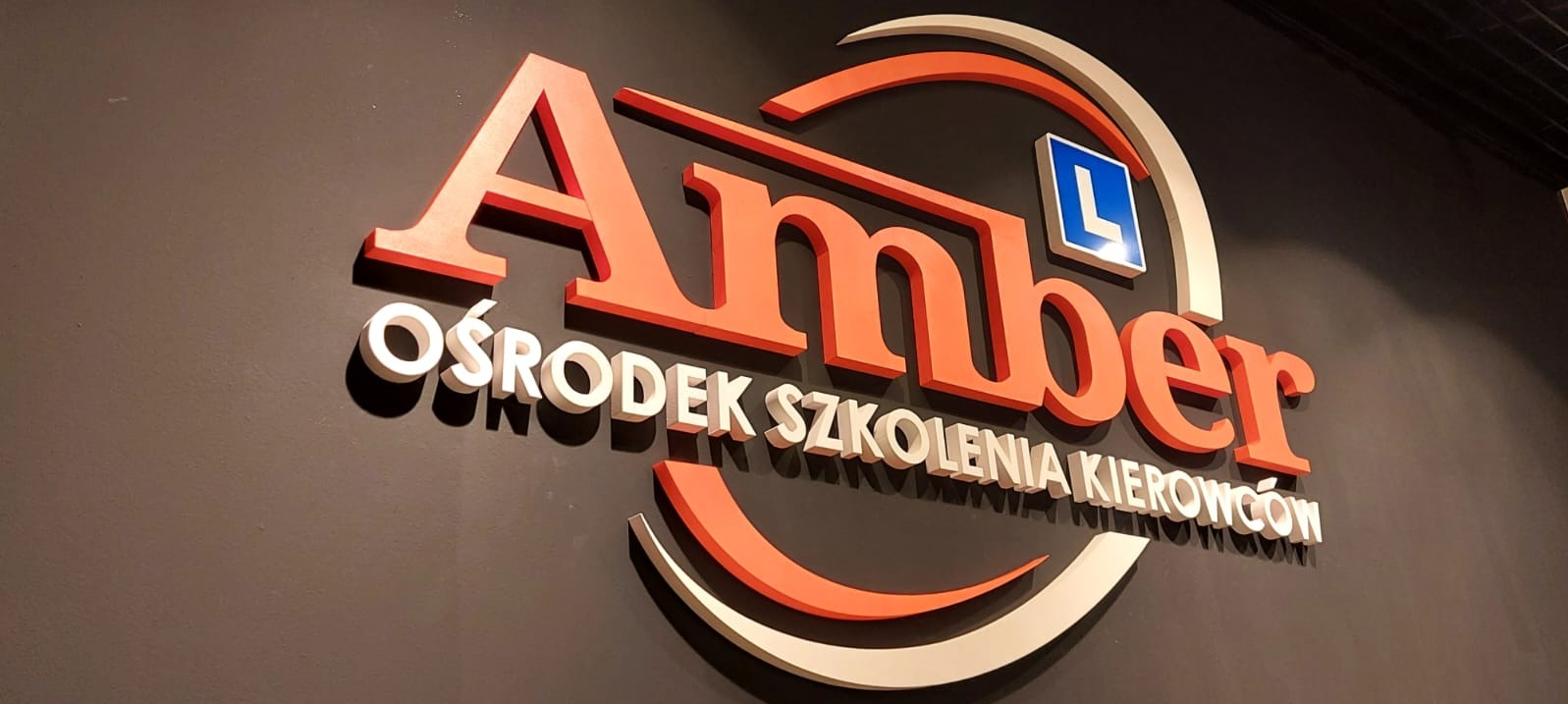 Logo szkoły jazdy 'Amber' z pomarańczowymi literami i symbolem 'L' na tle ciemnej ściany.
