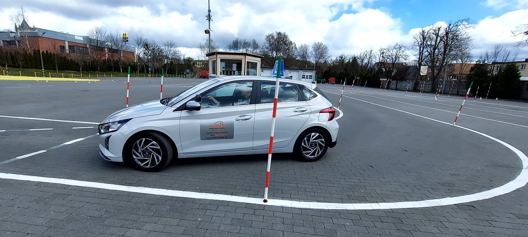 Srebrny samochód osobowy z naklejką firmy na karoserii, manewrujący na placu z pachołkami w trakcie egzaminu na prawo jazdy, widok z boku.