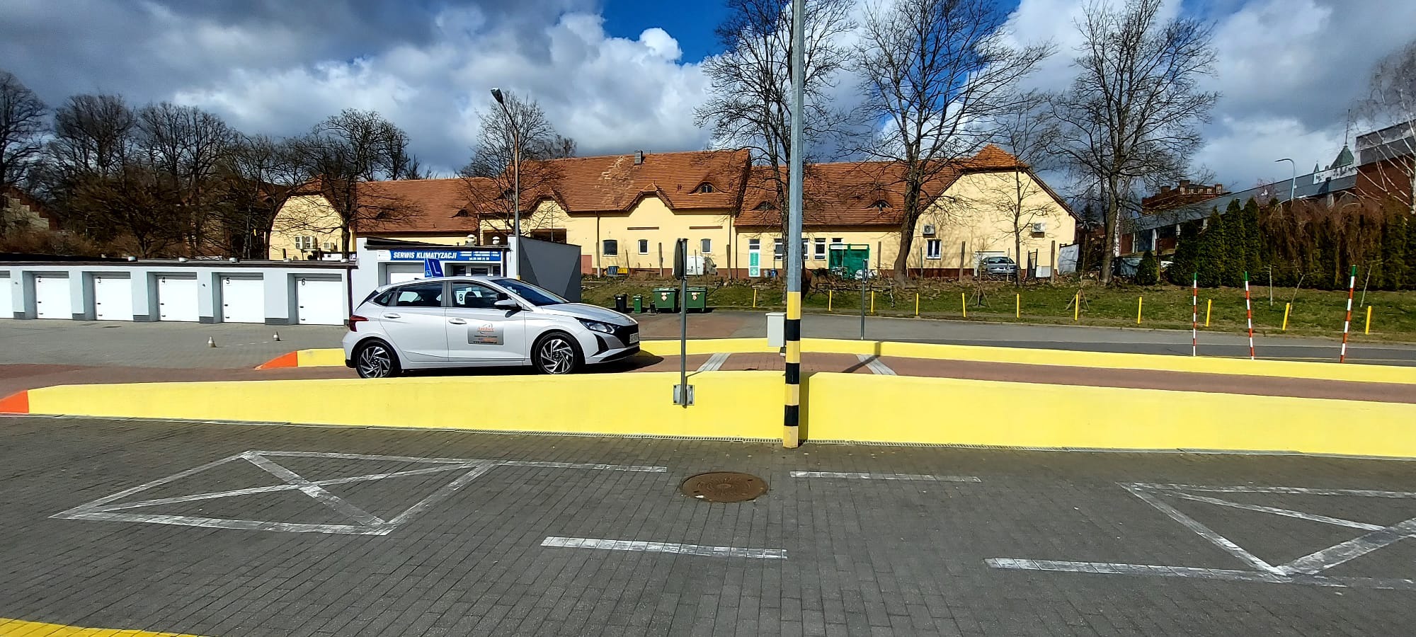 Srebrny samochód z naklejką szkoły jazdy na placu manewrowym z żółtą platformą, białymi liniami na kostce brukowej i budynkami w tle.