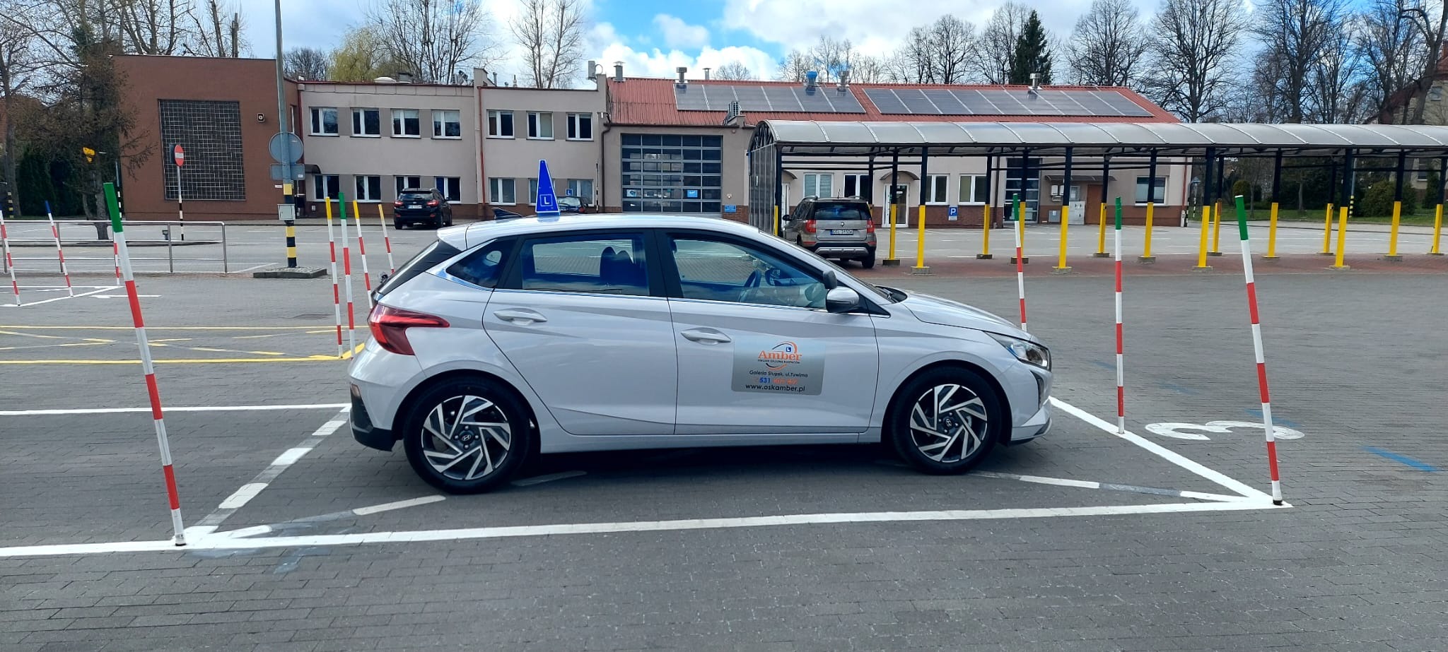 Szary samochód z niebieską literą 'L' na dachu, zaparkowany na placu manewrowym z pachołkami i liniami, budynek w tle.