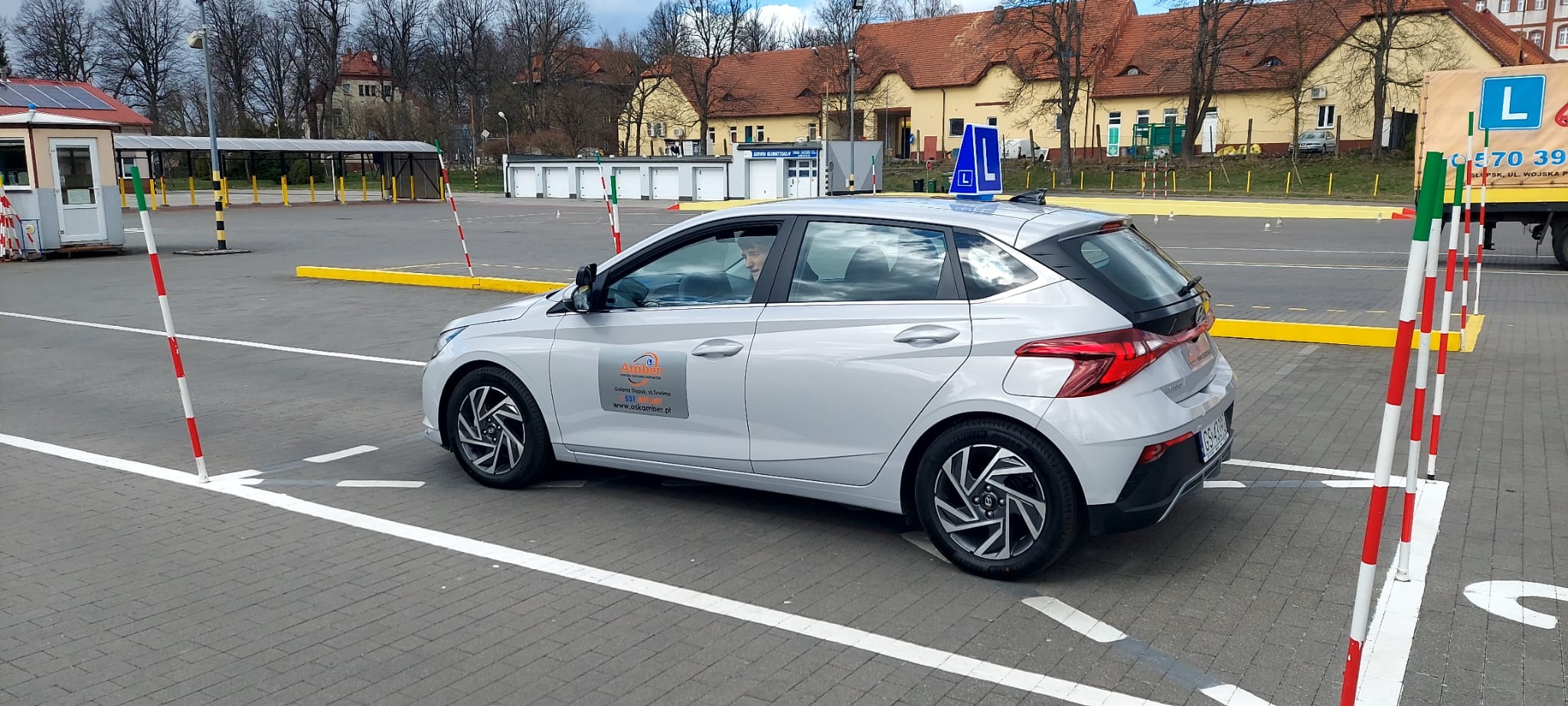 Srebrny samochód z literą 'L' na dachu, zaparkowany na placu manewrowym z pachołkami i liniami, widoczny kursant za kierownicą i logo firmy szkoleniowej na drzwiach