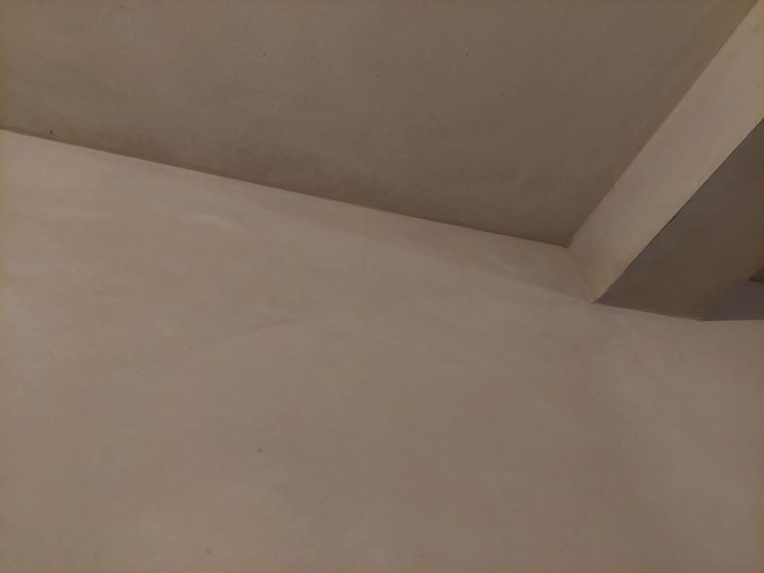 Gładka, jasna ściana po nałożeniu tynku, widok z dołu, z fragmentem sufitu i elementem konstrukcyjnym przy rogu. Delikatna faktura powierzchni.