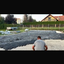 Prace ziemne - Pracownik w trakcie układania ciemnej kostki brukowej na podsypce żwirowej. Widoczny fragment ułożonej powierzchni, trawnik, ogrodzenie i budynek w tle.