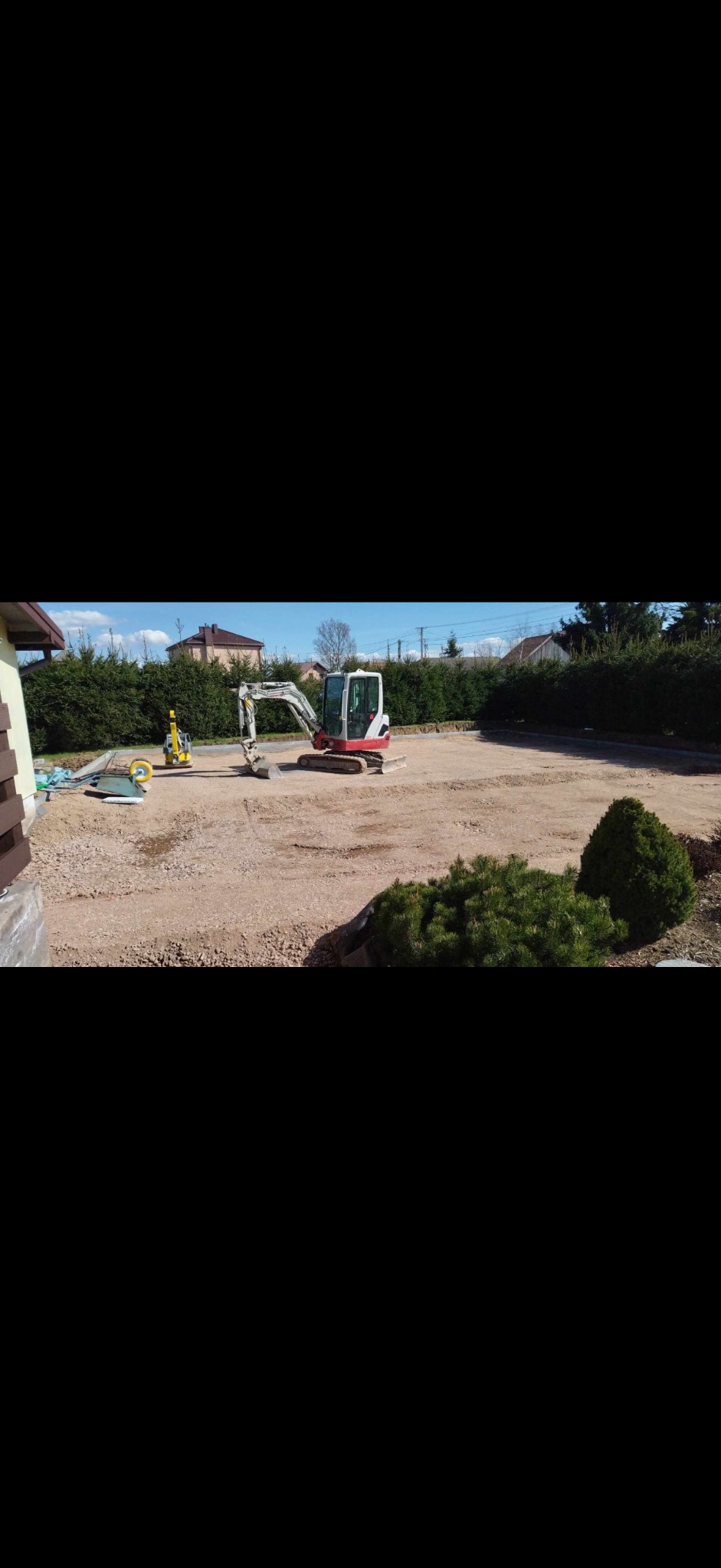 Mini koparka i walec na przygotowanym podłożu z kruszywa, prawdopodobnie pod kostkę brukową. Widoczne obrzeża i częściowo dom.