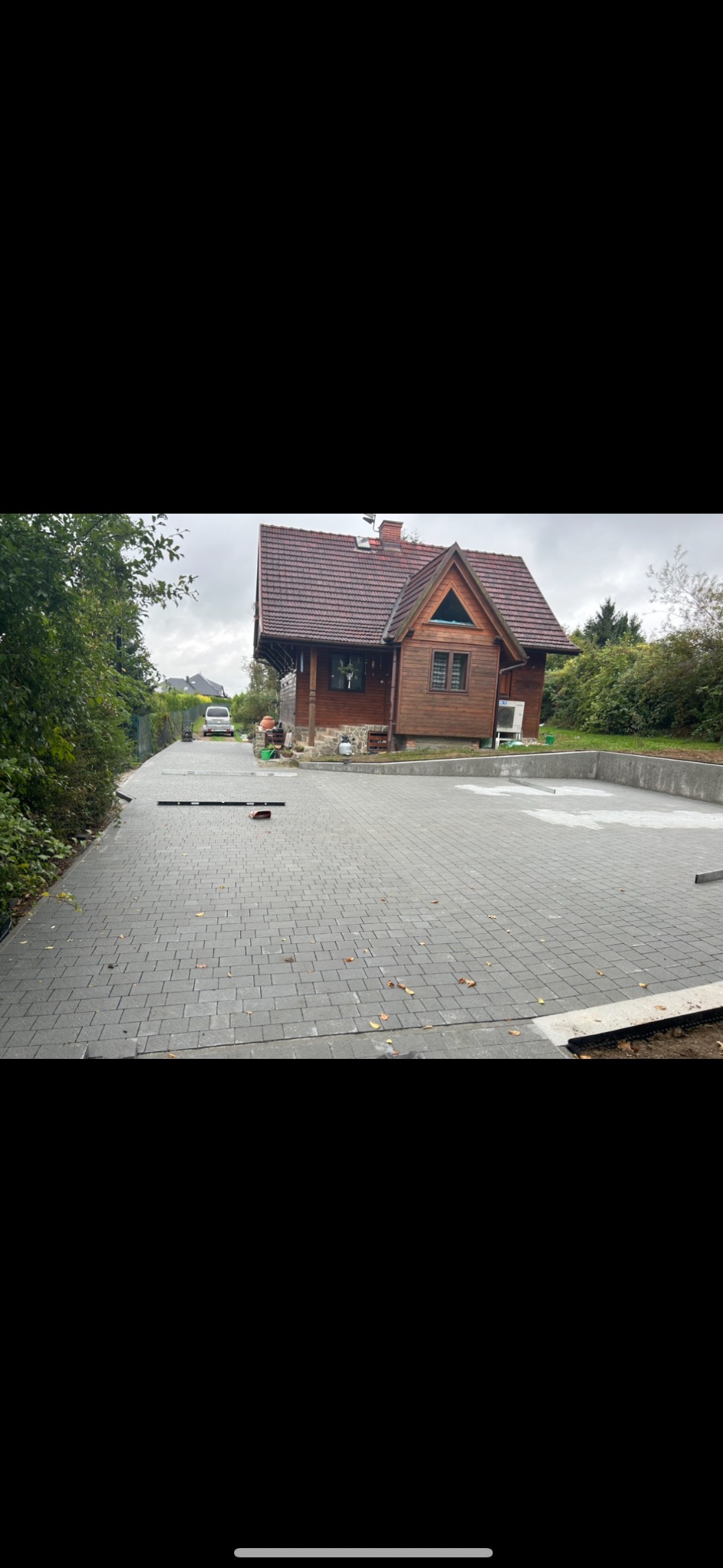 Ułożona, szara kostka brukowa na podjeździe do drewnianego domu z czerwoną dachówką. Widoczne narzędzia brukarskie i częściowo wykonane obrzeże. Tło: drzewa i pochmurne niebo.