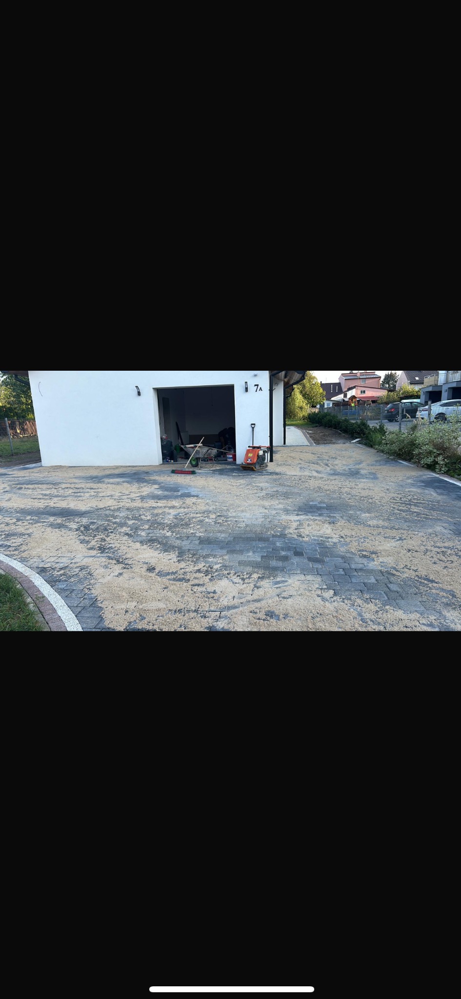 Ułożona kostka brukowa w odcieniach szarości przed budynkiem z numerem 7A, z widocznymi narzędziami i piaskiem wypełniającym szczeliny. Widok z perspektywy ulicy.