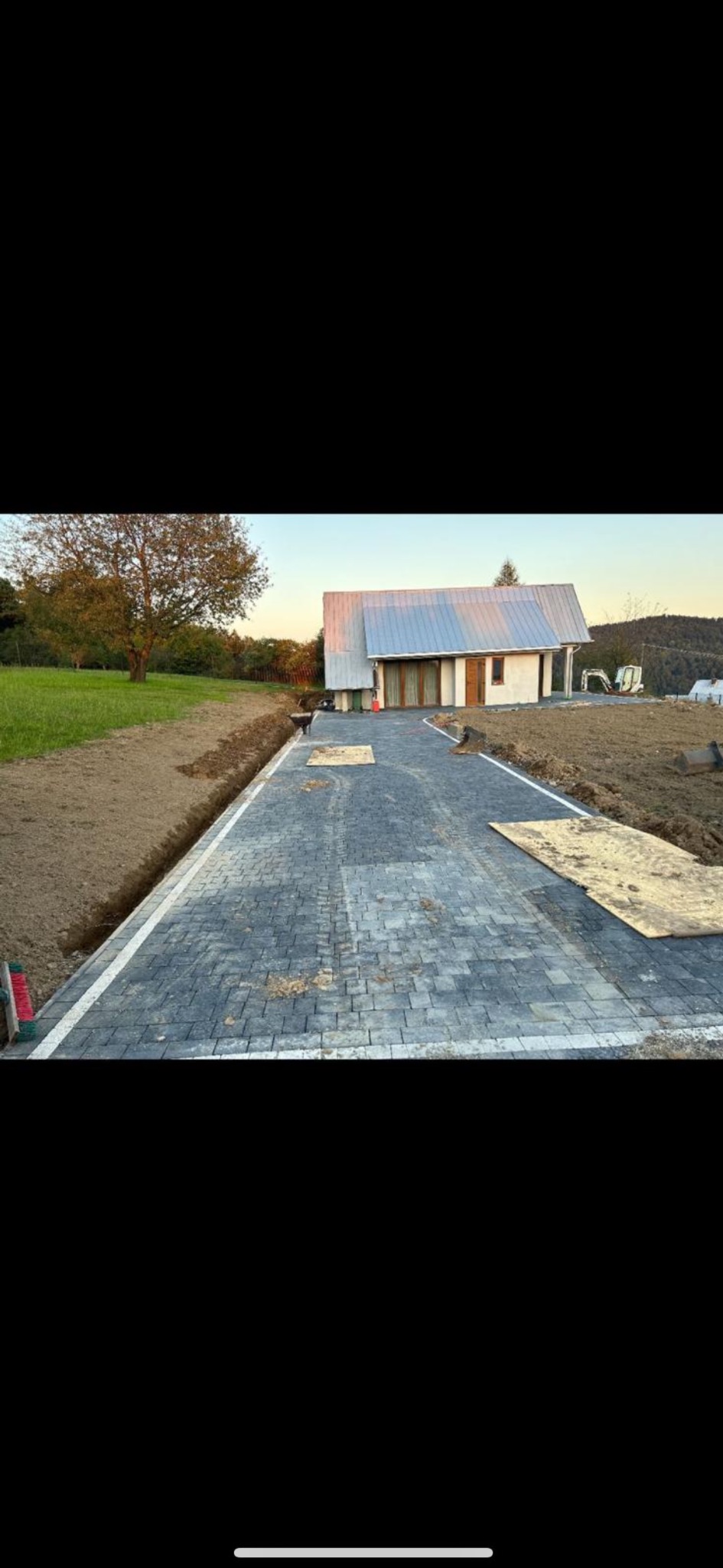 Ułożona, szara kostka brukowa prowadząca do domu z metalowym dachem. Widoczne obrzeża i przygotowany teren pod trawnik. Częściowo zakryta kostka płytami.