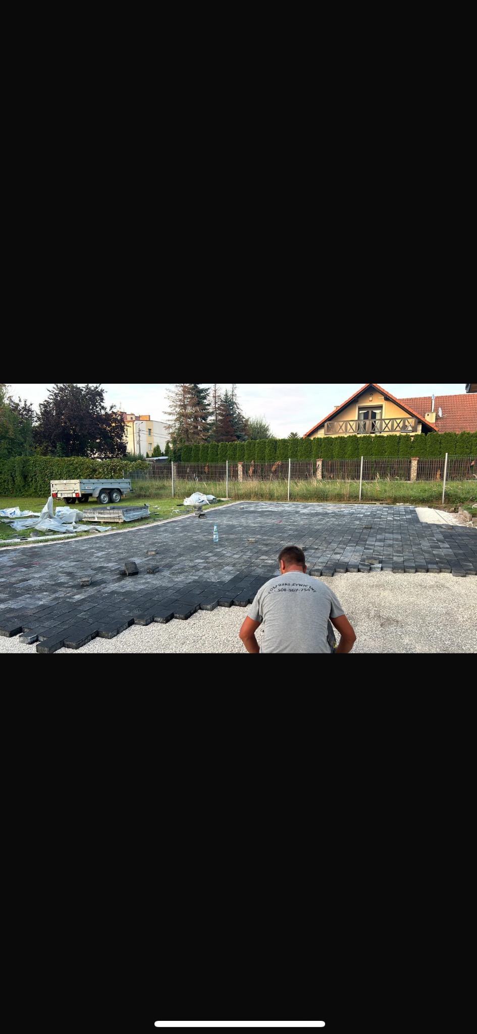 Pracownik w trakcie układania ciemnej kostki brukowej na podsypce żwirowej. Widoczny fragment ułożonej powierzchni, trawnik, ogrodzenie i budynek w tle.