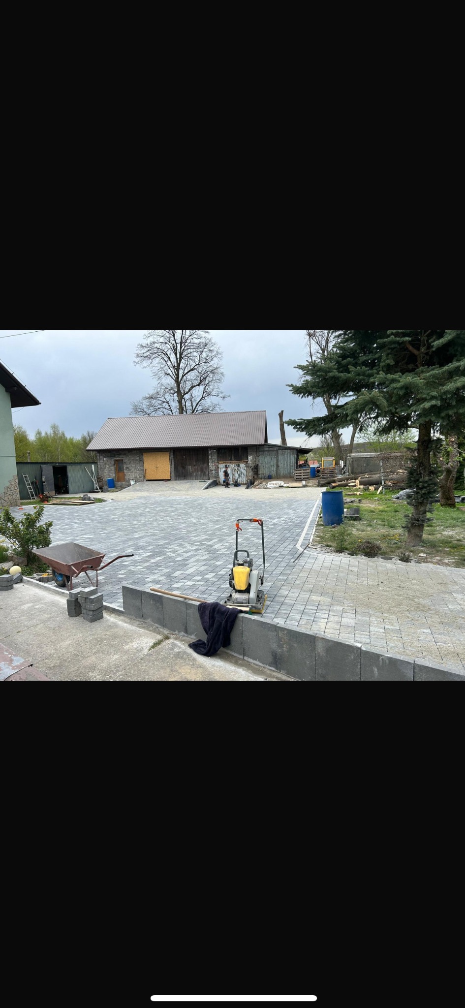 Ułożona kostka brukowa na posesji z wózkiem, ubijarką i budynkiem w tle. Widoczne świeżo ułożone obrzeża. Kostka w odcieniach szarości tworzy wzór.