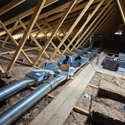 Hochlik sp. z o.o. - Dwie metalowe rury wentylacyjne w przestrzeni poddasza, prowadzone między drewnianymi belkami stropowymi, otoczone izolacją z wełny mineralnej. Widoczne narzędzia i materiały montażowe.