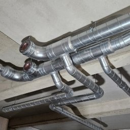 Hochlik sp. z o.o. - Srebrne, spiralne rury wentylacyjne zamontowane pod betonowym sufitem w surowym wnętrzu. Widoczne łączenia i mocowania.