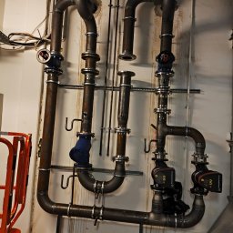 Hochlik sp. z o.o. - Złożona instalacja rur z zaworami i pompami Grundfos na ścianie. Widoczne spawane łączenia i elementy mocujące. Instalacja w pomieszczeniu technicznym.
