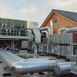 Hochlik sp. z o.o. - Rozbudowana instalacja wentylacyjna na dachu budynku z drewnianą konstrukcją. Widoczne liczne rury, kanały wentylacyjne i urządzenia. Elementy instalacji w kolorze srebrnym i zielonym.